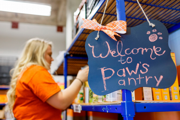 Women stocks shelves with canned goods, sign in the foreground says Welcome to Paw's Pantry.